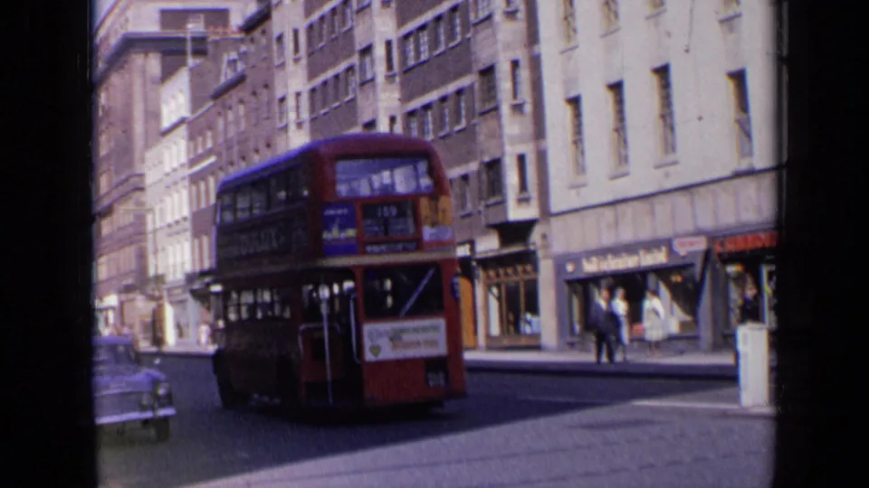 Wide 1963 London street view with a red double-decker bus driving away, pedestrians on the pavement and rows of shopfronts and buildings.