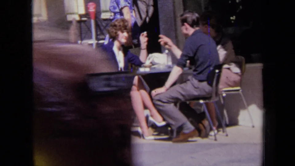 A passerby briefly crosses the 1963 London sidewalk scene as the seated pair continue their conversation.