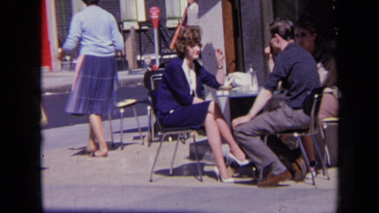 Couple seated at a sunny outdoor cafe table with pedestrians behind them.