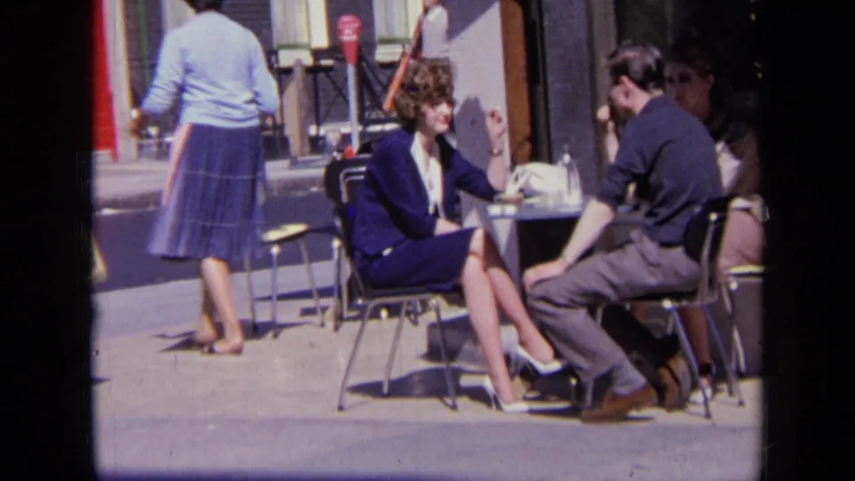 In 1963 London, a woman in a blue suit and a man in dark clothing sit talking at a pavement cafe table.
