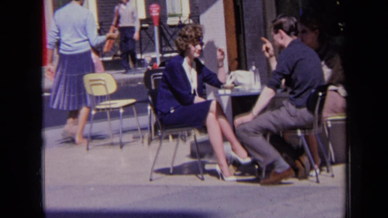 Outdoor cafe conversation with a woman gesturing and people walking behind.