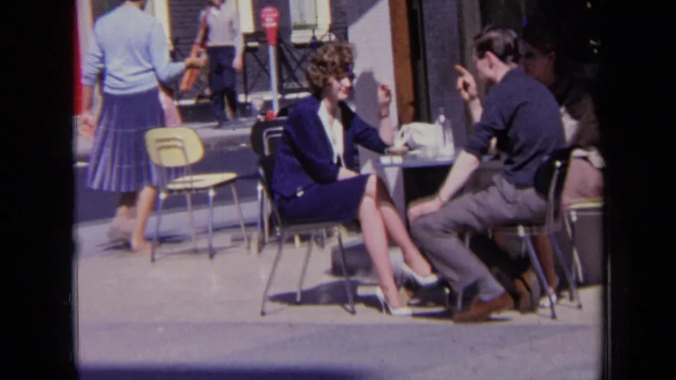 The London cafe scene continues as the seated woman gestures while speaking with the man across the small table.