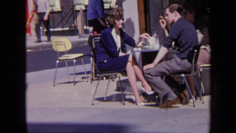 A seated couple continues their cafe conversation, with a yellow chair and passing street activity visible in the London background.