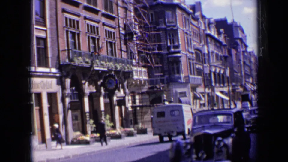 1963 London street view with multi-story facades, sidewalk pedestrians, a white delivery van, and a dark car in the road.