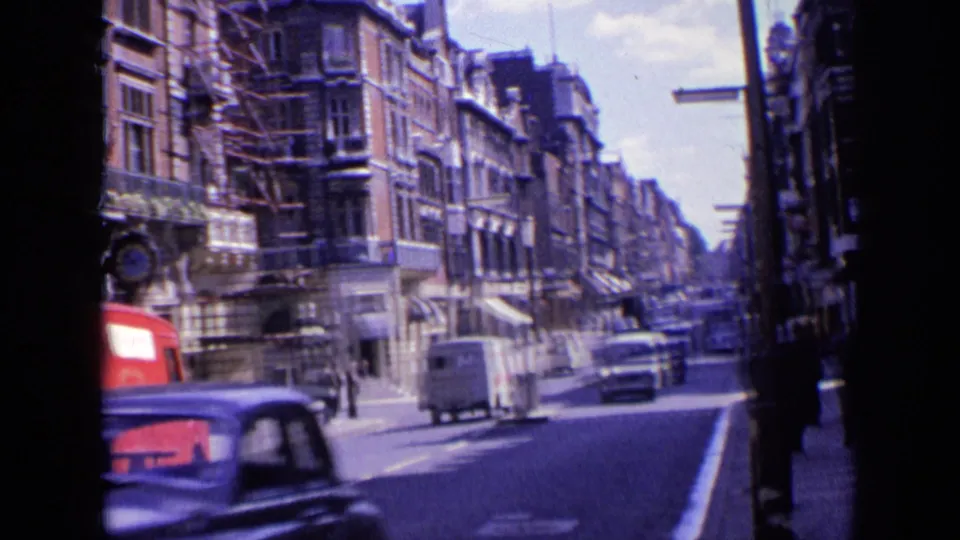 Forward-moving 1963 London street view with a red van at left, a white van ahead, cars, pedestrians, and dense urban facades.