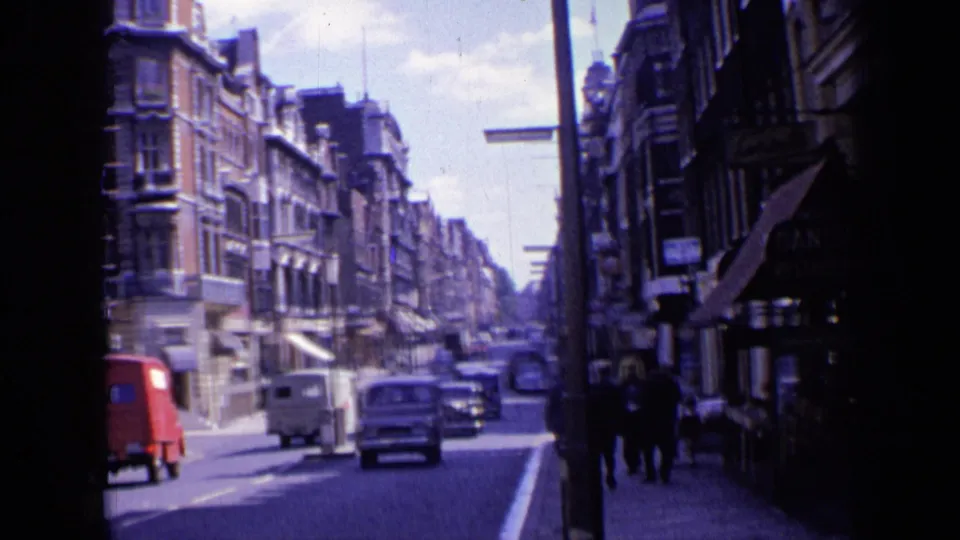 1963 London street scene with a red van, white van, several cars, pedestrians on the sidewalk, and shopfront awnings along older buildings.