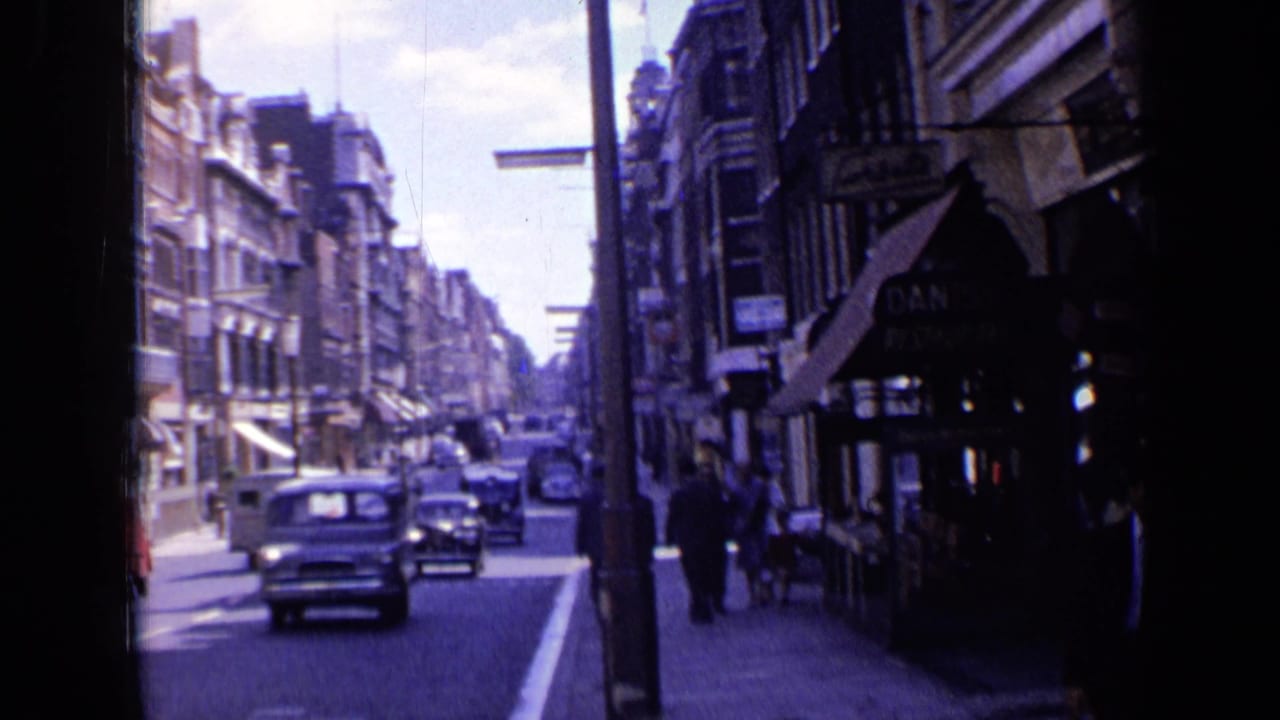 1963 London street view from a moving vehicle, showing mid-century cars, pedestrians, brick facades, and storefront awnings.