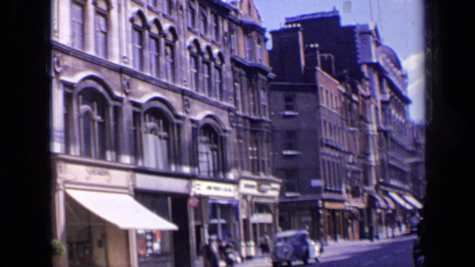 1963 London street view with ornate brick facades, storefront awnings, pedestrians, and a mid-century car near the curb.