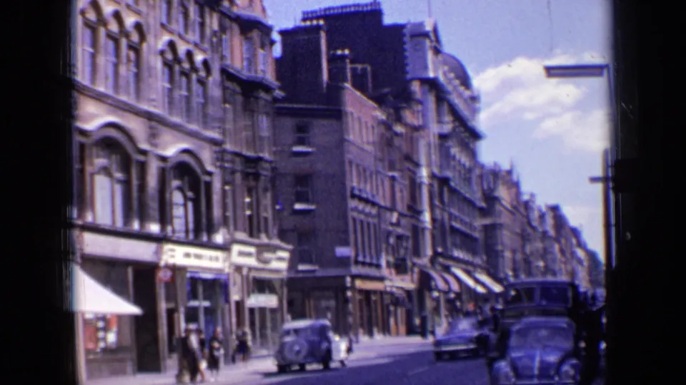 1963 London street corner with shop signs, pedestrians, a parked mid-century car, and another car passing close to the camera.