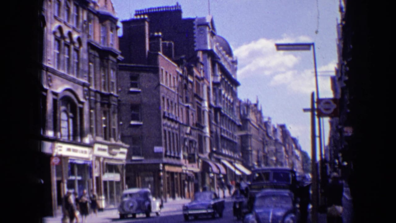 London street with vintage cars, pedestrians, shopfronts, and tall buildings in sunlight.