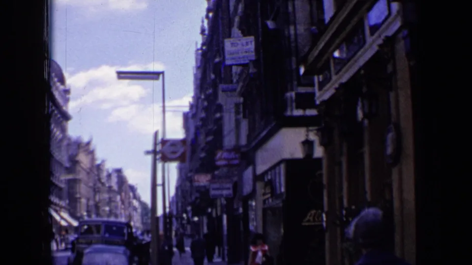 1963 London street scene showing shop signage, pedestrians, vintage cars, an Underground roundel, and dense commercial buildings.
