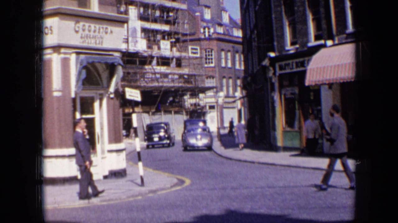 London street corner with pedestrians, storefront signs, scaffolding, and vintage cars on a narrow road.