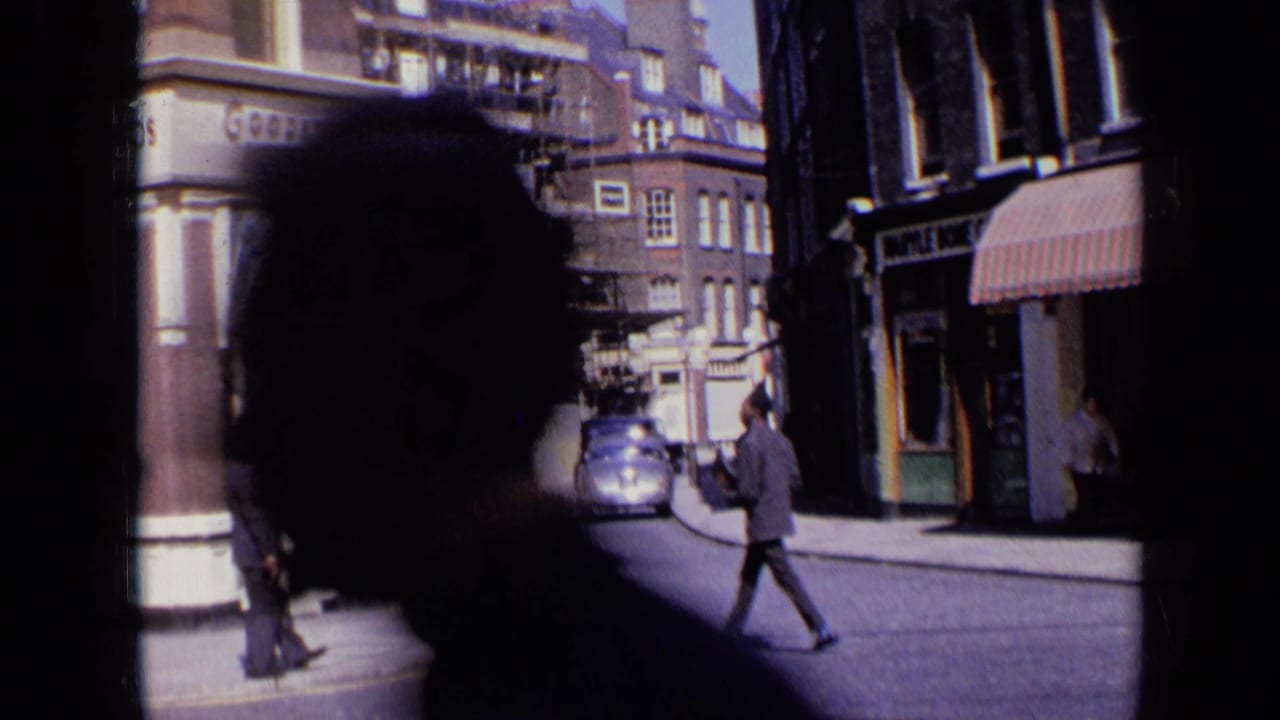 1963 London street scene partly blocked by a close foreground silhouette, with a vintage car, pedestrians, and storefronts still visible beyond.