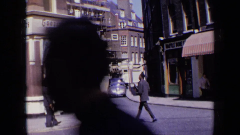 1963 London street scene partly blocked by a close foreground silhouette, with a vintage car, pedestrians, and storefronts still visible beyond.