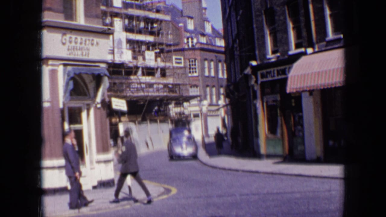 1963 London street corner with a man standing near a shop entrance, another pedestrian crossing, scaffolding above, and a vintage car on the road.