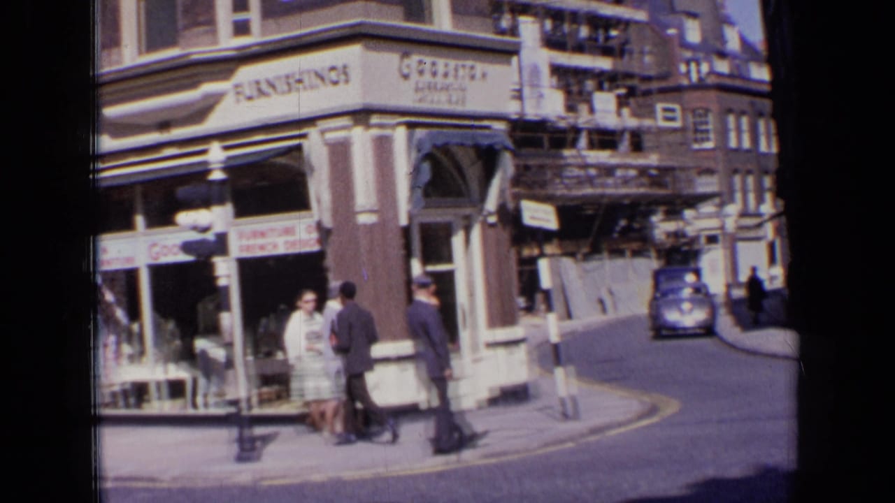 1963 London corner view showing a Goodison shopfront, pedestrians near the curb, directional street signs, scaffolding, and a blue vintage car driving past.