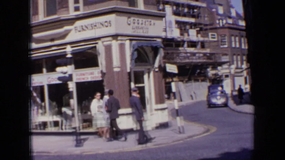 1963 London corner view showing a Goodison shopfront, pedestrians near the curb, directional street signs, scaffolding, and a blue vintage car driving past.