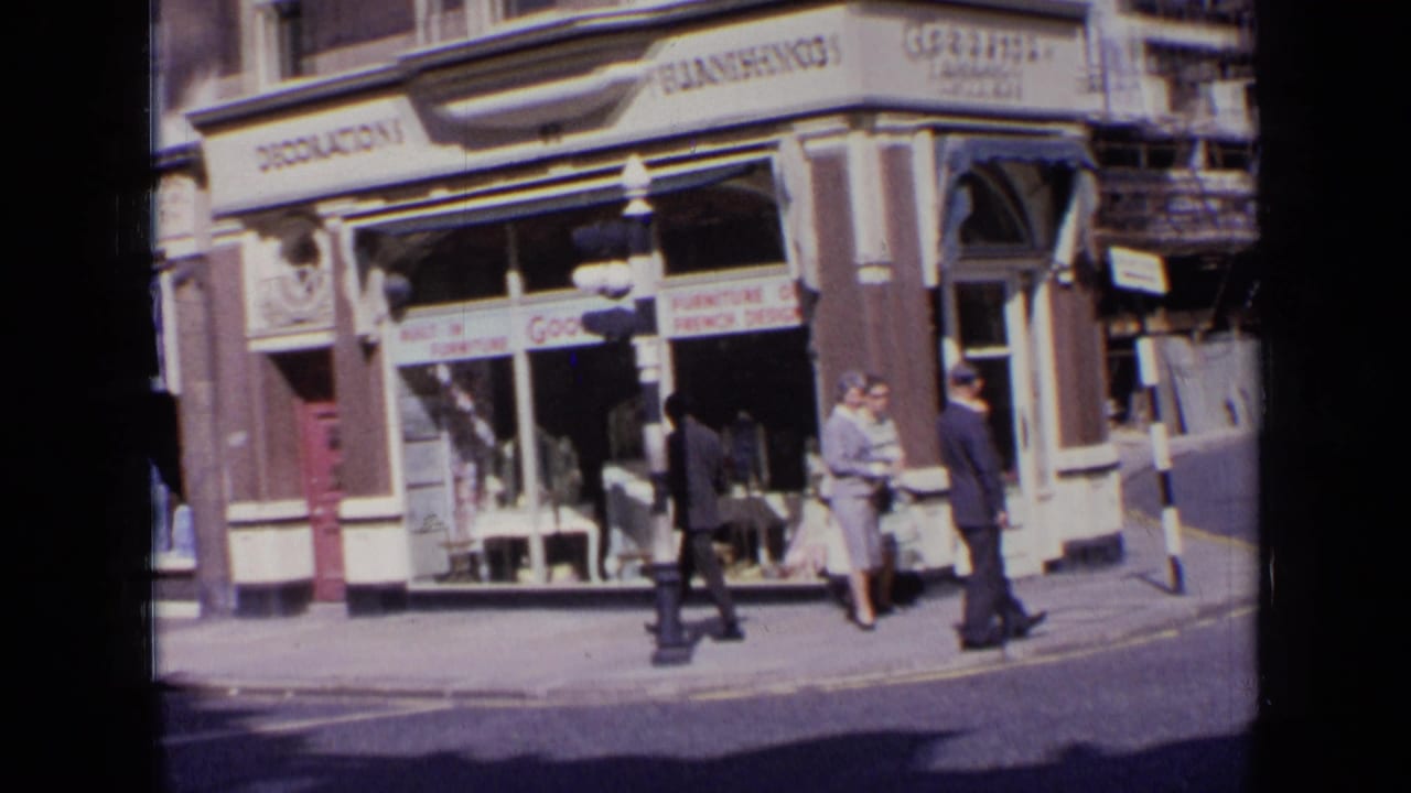 1963 London street view with suited pedestrians walking past shop windows and visible retail signs in 8mm home-movie footage.