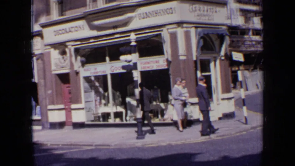 1963 London street view with suited pedestrians walking past shop windows and visible retail signs in 8mm home-movie footage.