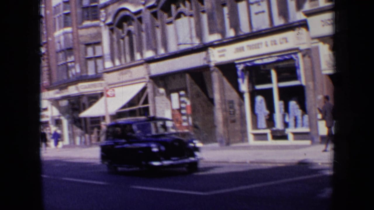 A dark period car moves through the foreground while London shopfronts and pedestrians remain visible behind it.