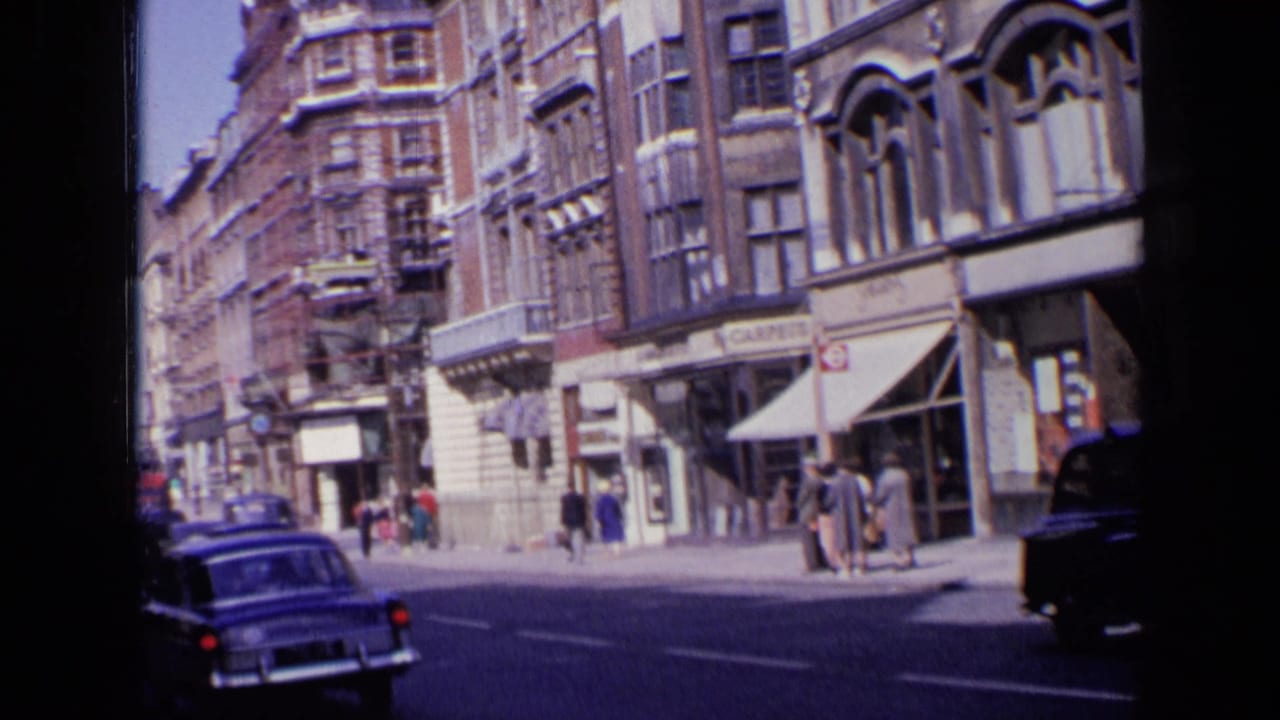 Wider 1963 London street view showing multi-story buildings, shop awnings, pedestrians, and period traffic in home-movie color.