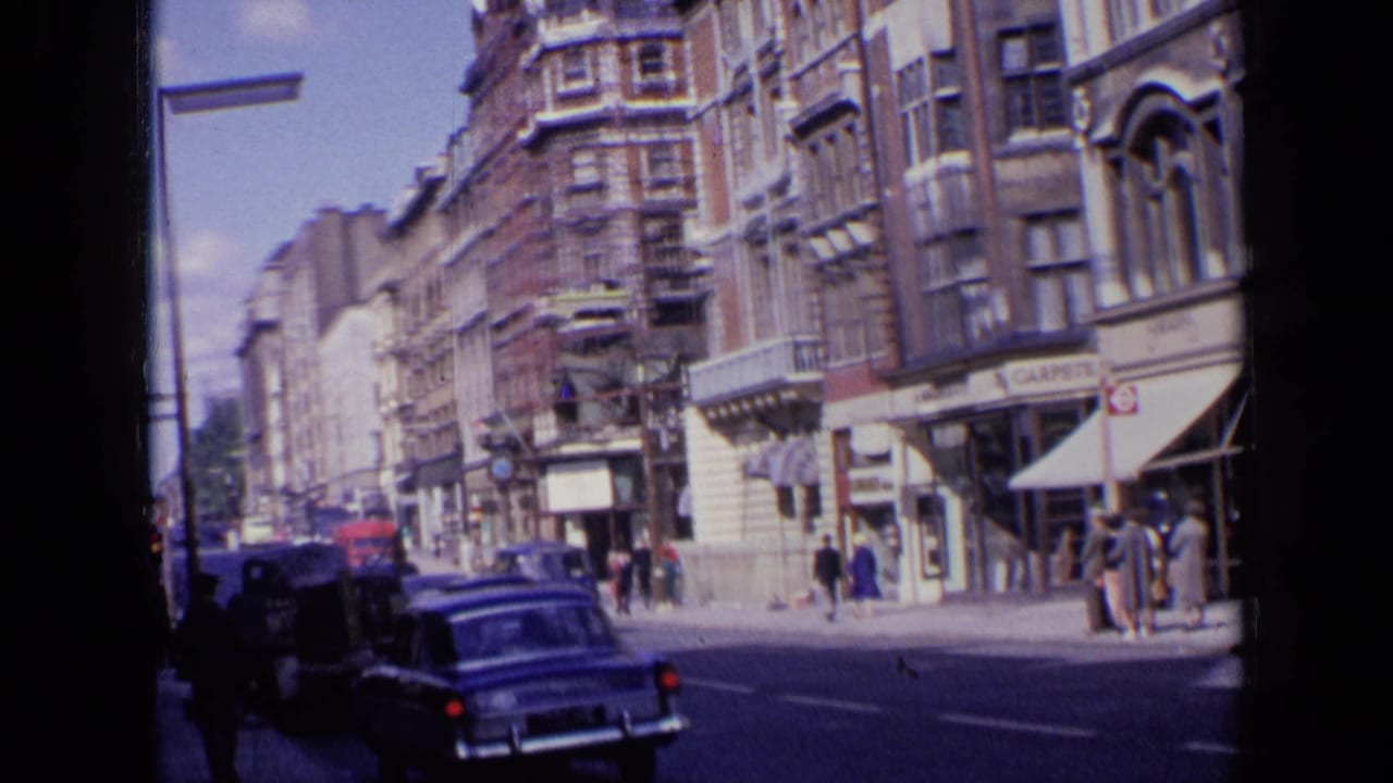 1963 London street view with mid-century cars, brick commercial buildings, pedestrians, awnings, and a red double-decker bus farther down the road.
