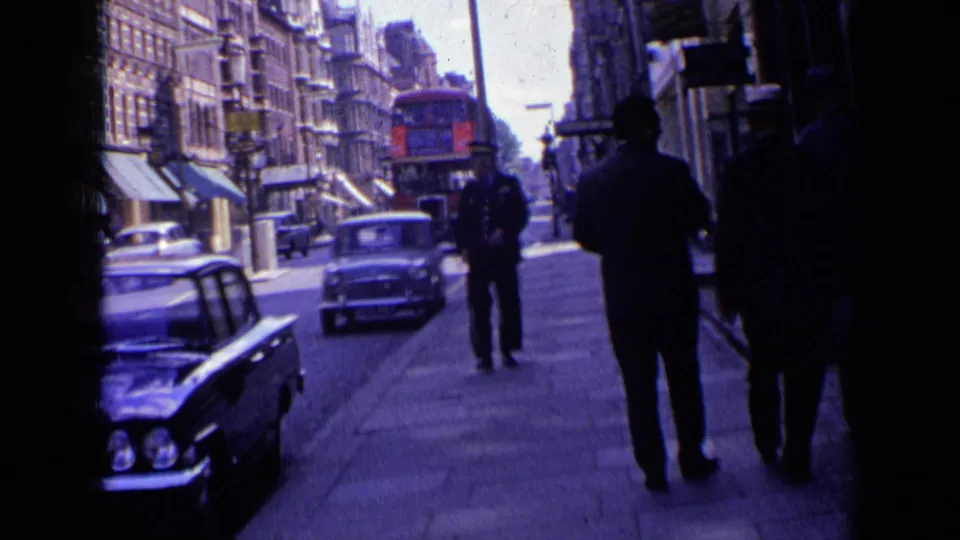 1963 London sidewalk view with a uniformed police officer, pedestrians, mid-century cars, shopfront awnings, and a red double-decker bus in traffic.