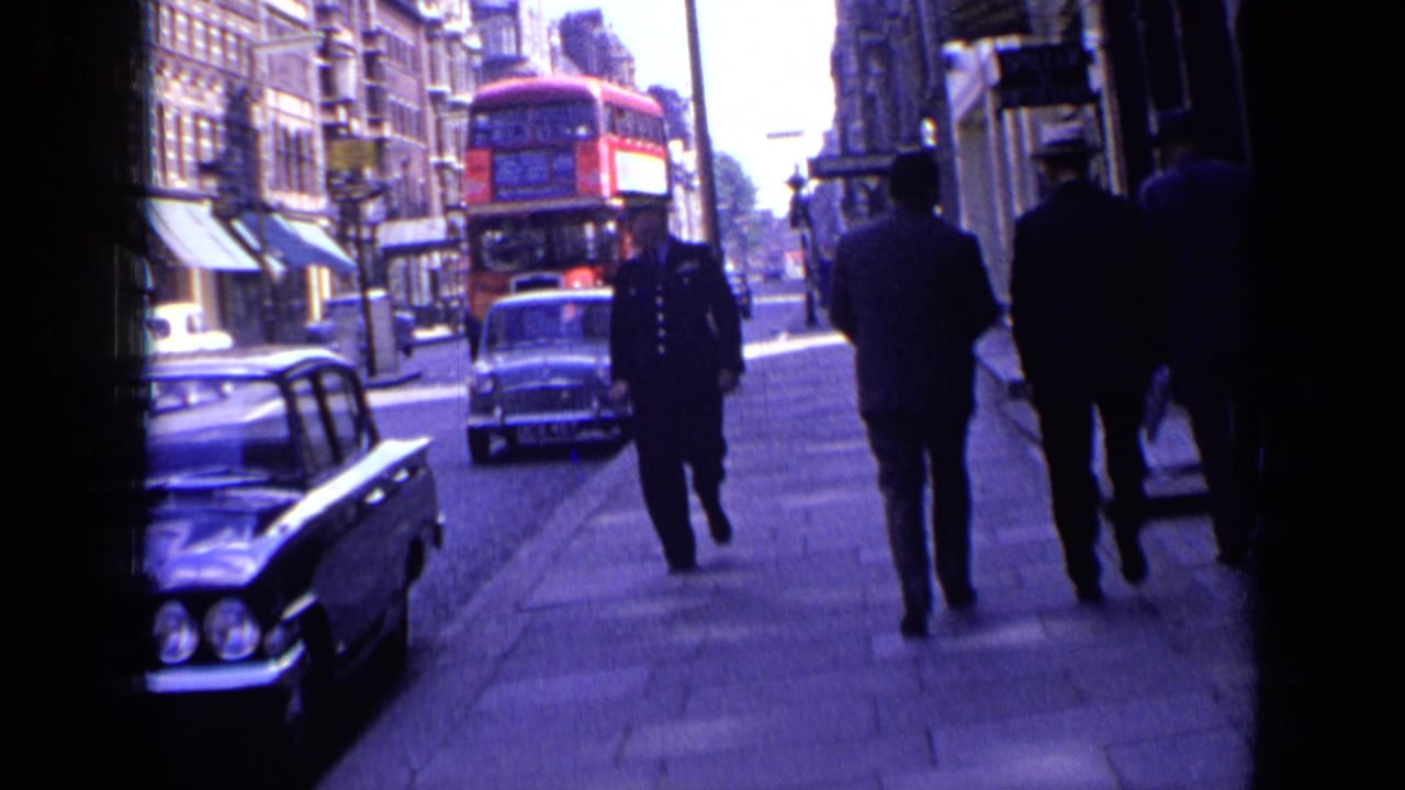 1963 London street scene with a uniformed police officer approaching the camera, pedestrians on the sidewalk, classic cars, and a red bus behind.