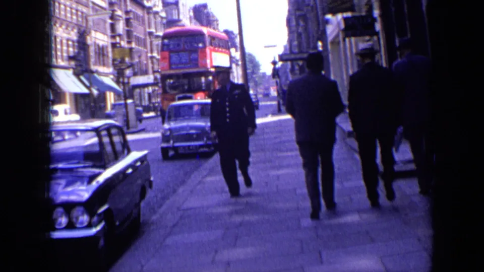 1963 London street scene with a uniformed police officer approaching the camera, pedestrians on the sidewalk, classic cars, and a red bus behind.