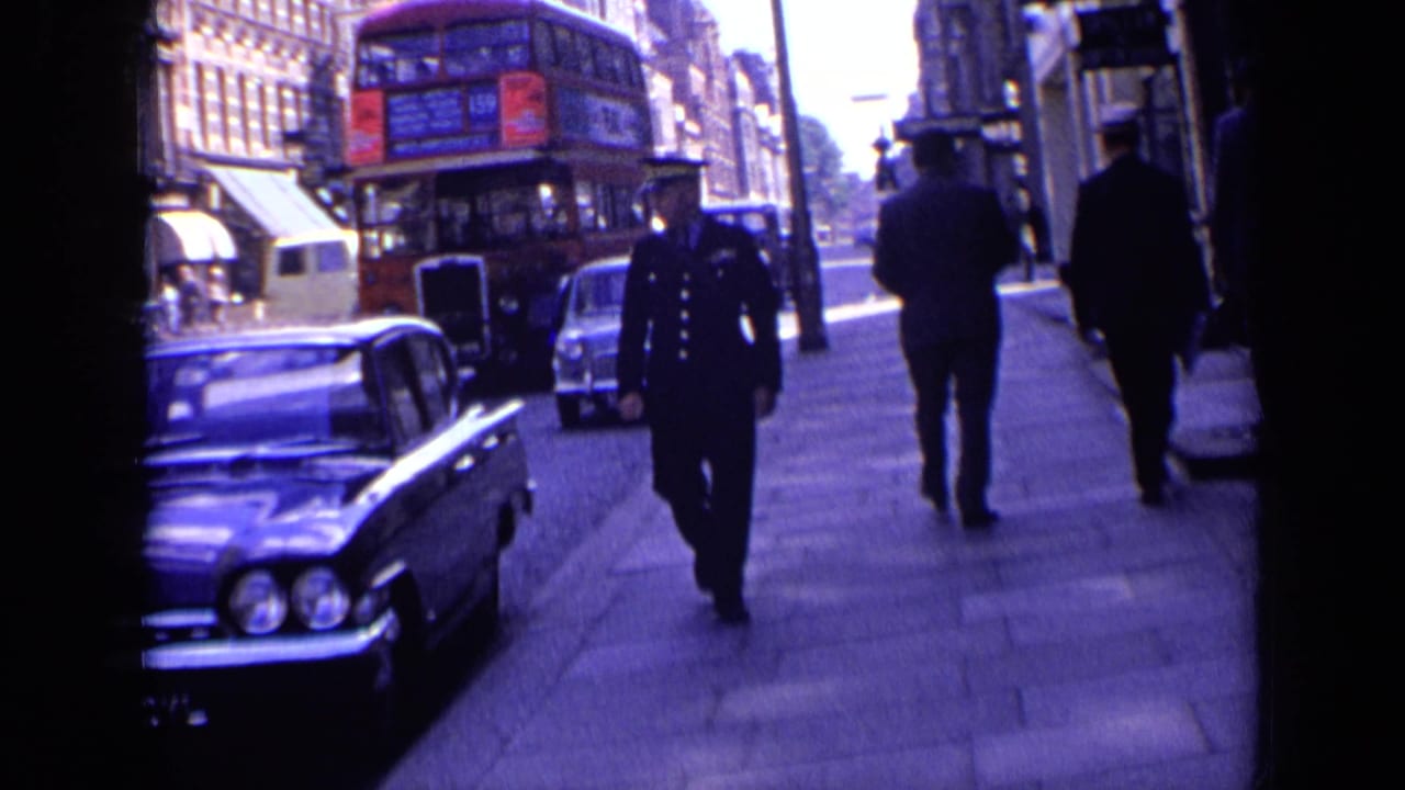 1963 London sidewalk and roadway view with a red double-decker bus, uniformed police officer, pedestrians, classic cars, and brick shopfront buildings.