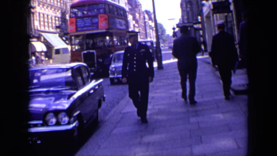 1963 London sidewalk and roadway view with a red double-decker bus, uniformed police officer, pedestrians, classic cars, and brick shopfront buildings.