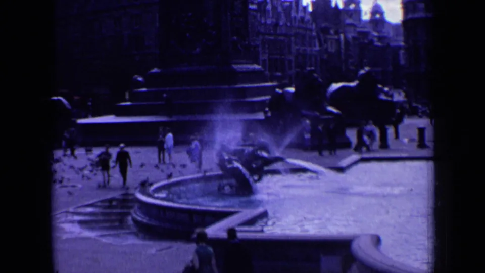 1963 London home-movie view of Trafalgar Square with a fountain spraying, pedestrians walking, and surrounding city architecture.