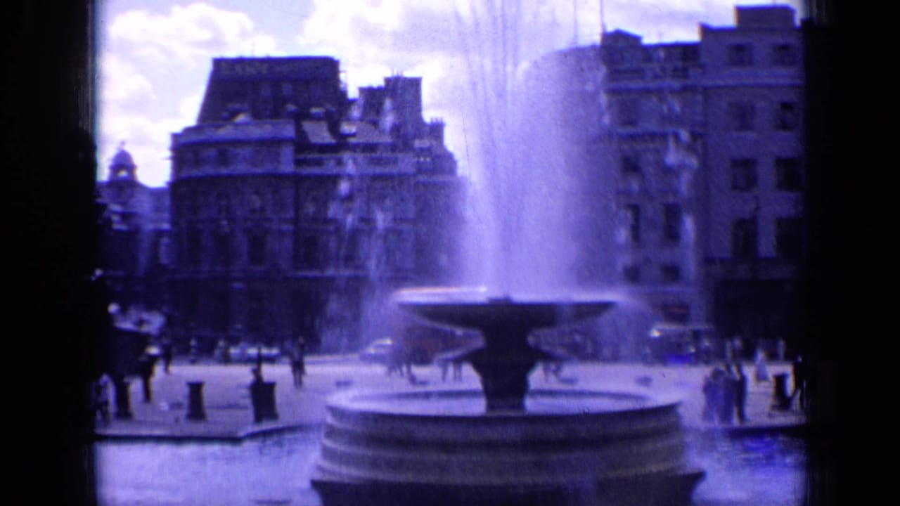 Wide 1963 8mm frame of a London square, with the fountain centered and pedestrians crossing near surrounding buildings.
