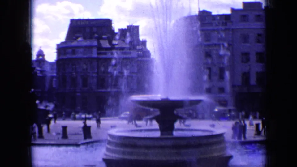 Wide 1963 8mm frame of a London square, with the fountain centered and pedestrians crossing near surrounding buildings.