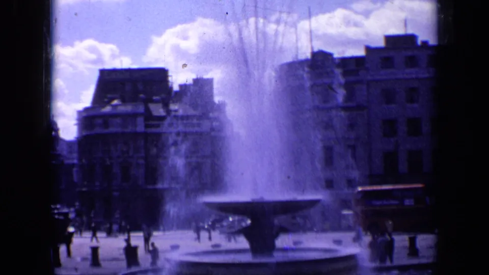1963 London 8mm frame showing a public square fountain, surrounding pedestrians, and city buildings under a bright sky.