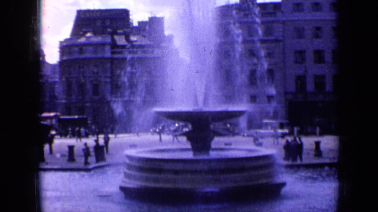 Closer view of a large fountain with water spray, pedestrians, and buildings in the background.