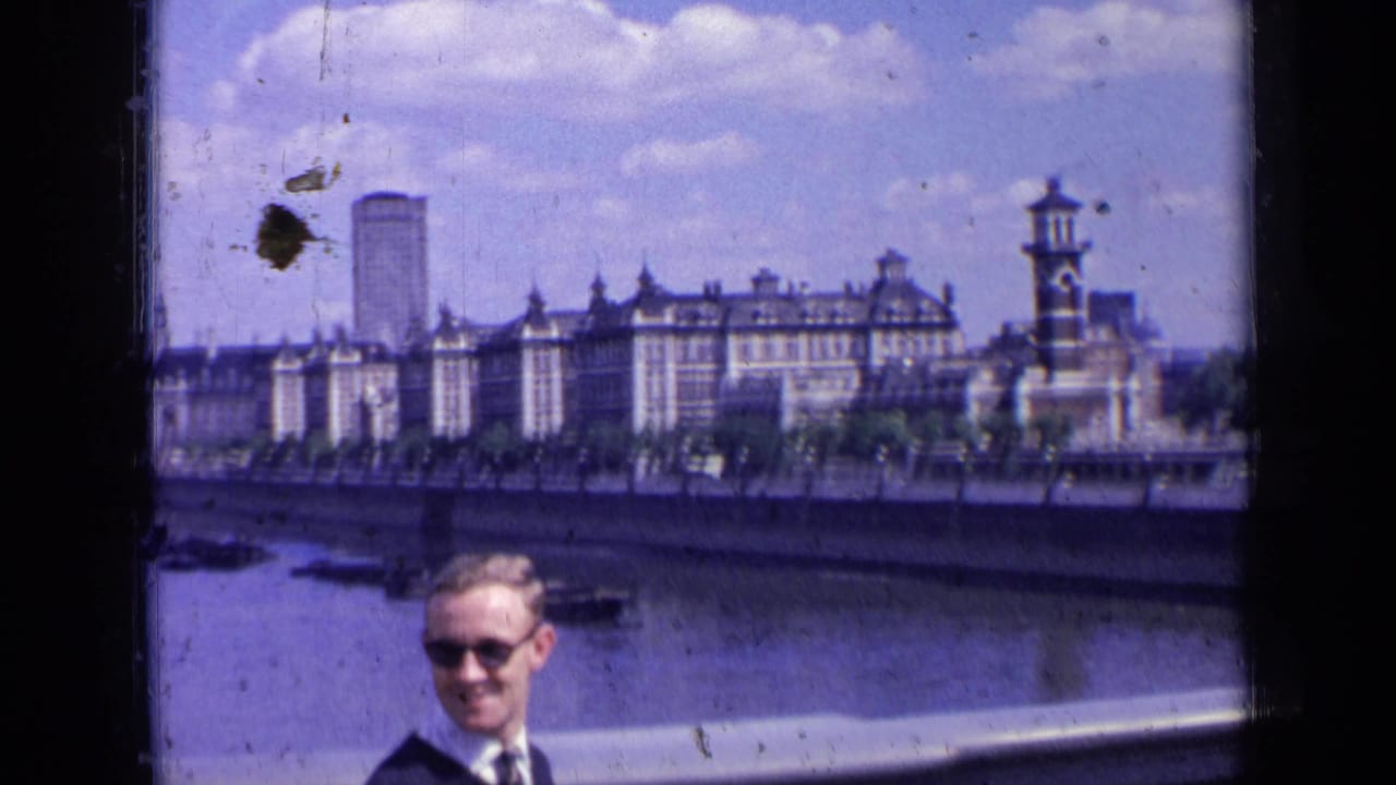 1963 London home-movie frame showing a smiling man by the Thames, with riverside buildings and a tower-like structure behind him.