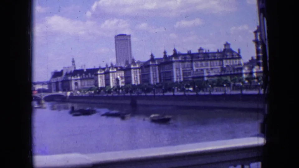 1963 London riverside view during a pan, showing boats on the river, a bridge arch at left, waterfront buildings, and a tall background tower.