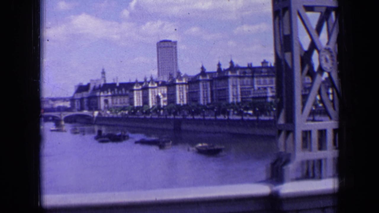 1963 London riverfront seen past a bridge structure, with small boats on the water, long buildings along the bank, and a tall tower behind them.