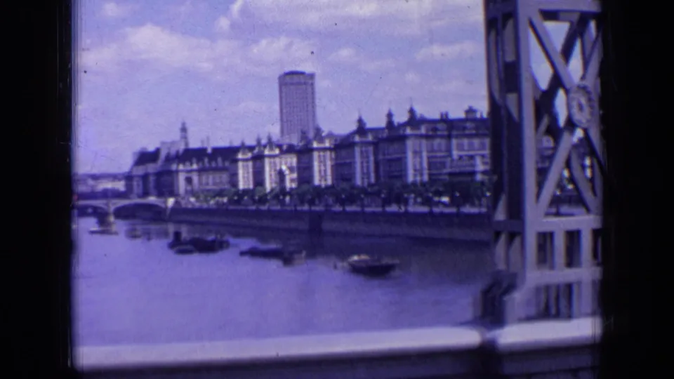 1963 London riverfront seen past a bridge structure, with small boats on the water, long buildings along the bank, and a tall tower behind them.