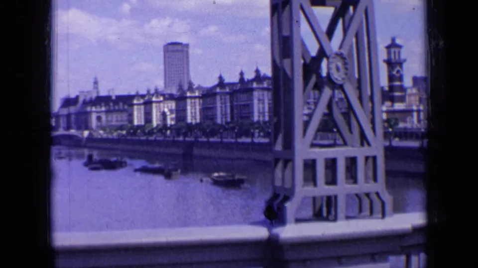 1963 London 8mm frame looking from a boat past bridge metalwork toward riverside buildings, boats, and a tall tower.