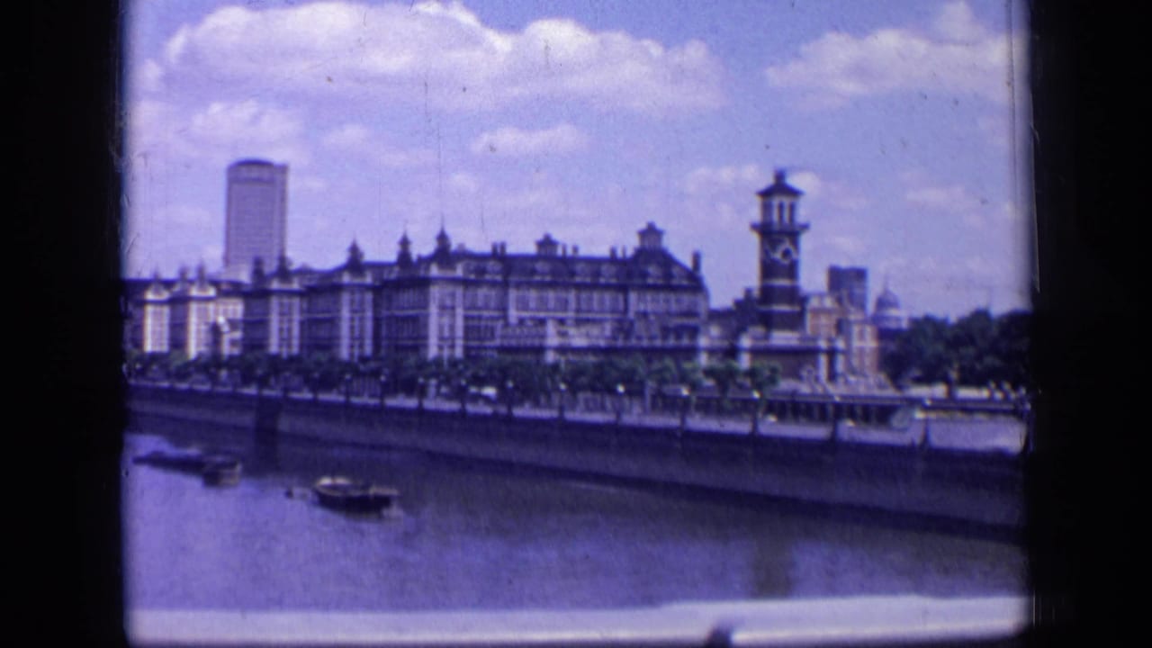 1963 London 8mm view across the river toward a long embankment building, a clock-tower-like structure, boats, and a tall tower.