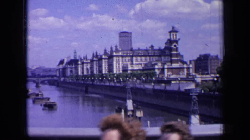 1963 London riverfront view with the grand building, boats, distant bridge, and the tops of people briefly entering the foreground.