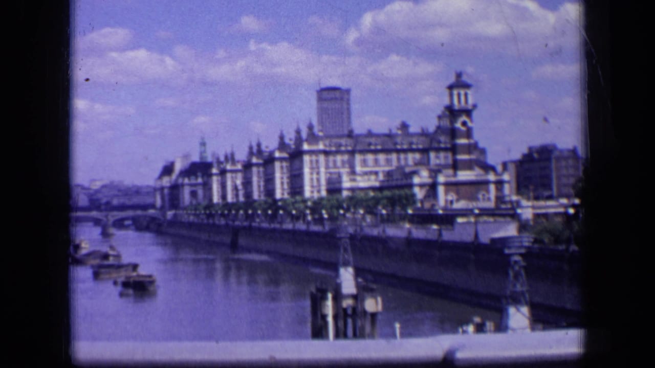 1963 London home-movie frame of a large riverside building with tower details, riverboats, a distant bridge, and soft 8mm color.