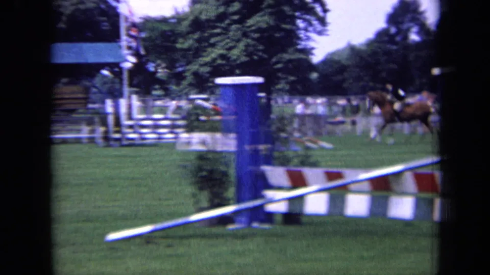The home-movie frame shows a rider and horse beyond a blue jump post and red-and-white rail at a London equestrian venue in 1963.