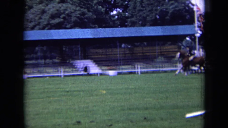 A rider on horseback moves along the edge of a grassy 1963 London equestrian course near a covered stand and low fencing.