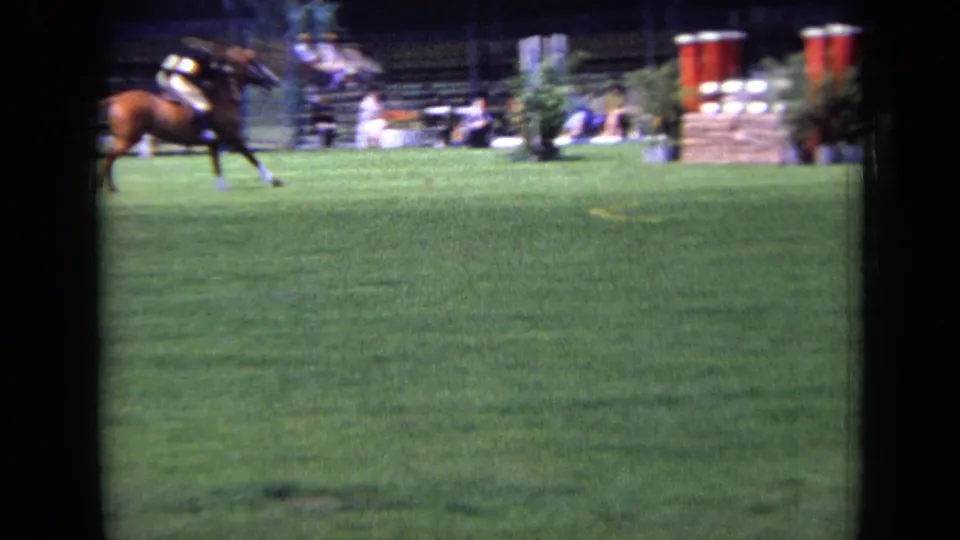 A horse and rider move quickly across the grass arena in London, with red and white jumping equipment and spectators in the background.