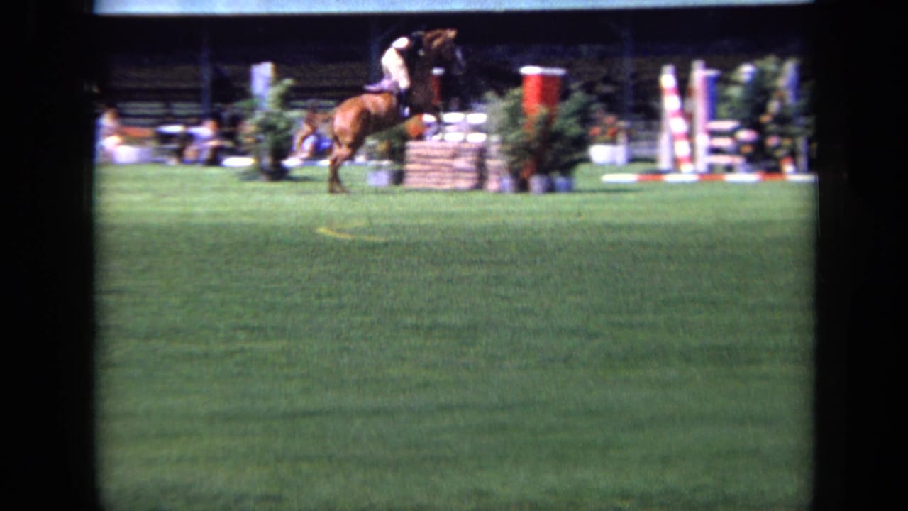 In supported 1963 London home-movie footage, a rider on a brown horse clears a jump in a grassy equestrian arena.