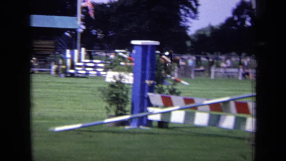 A blue-and-striped jump dominates the foreground while the horse and rider pass farther back on the grass course.