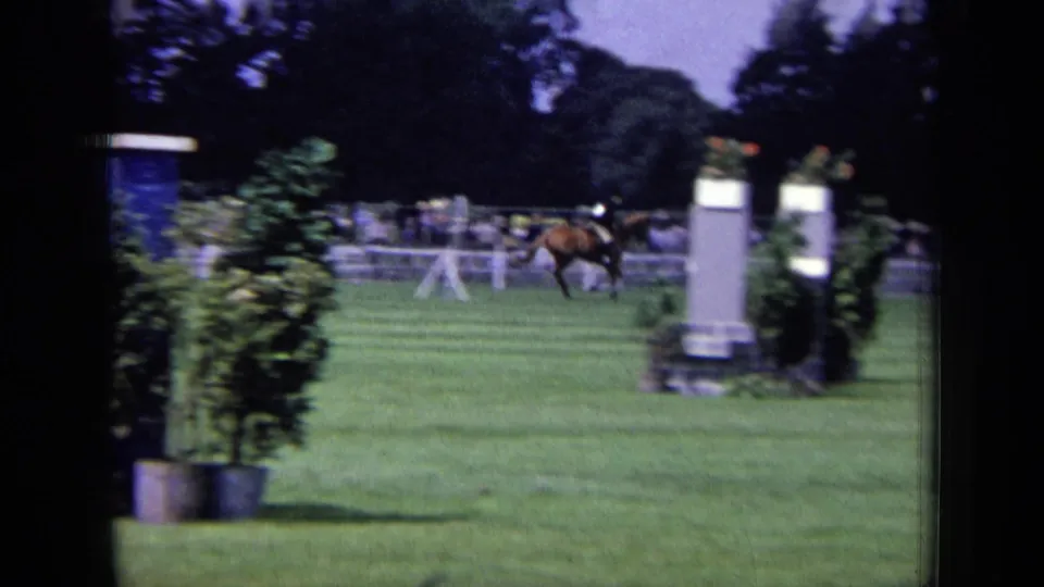 1963 London 8mm frame of a mounted rider crossing the field between equestrian obstacles and boundary fencing.